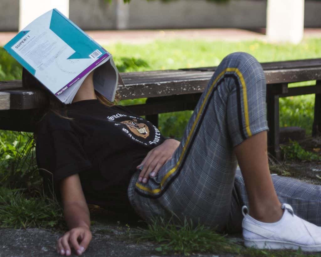 Teenager with a book covering her face while she rests her head against a bench